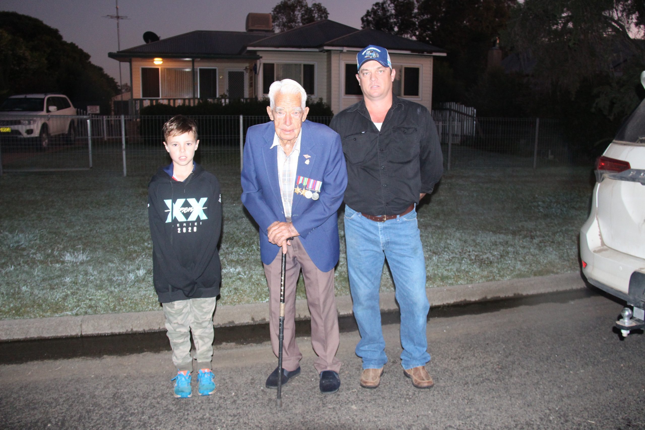 John Collett, centre, with his great-grandson Nate Dewson and grandson Luke Dewson marking a very different Anzac Day in 2020 listening to Peter Carrett playing the Last Post on his driveway.