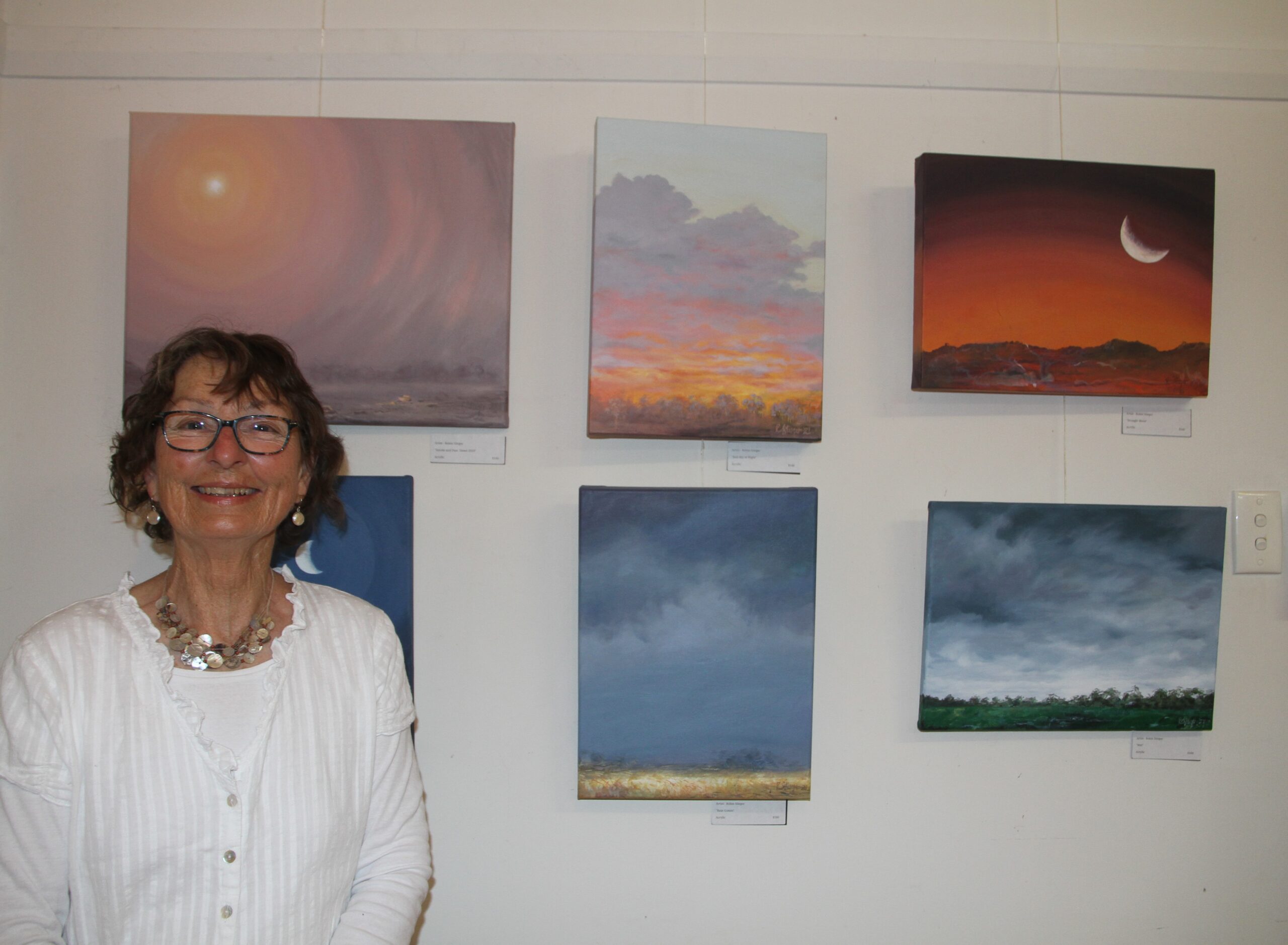 Robin Stieger with her exhibits, top from left, ‘Smoke and Dust. Dawn 2019’, ‘Red Sky at Night’, ‘Drought Moon’, bottom from left, ‘Tipping Moon’, ‘Rain Comin’ and ‘Wet’, all in acrylics.